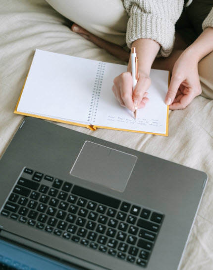 person writing on a book in front of a computer - Photo by Ivan Samkov from Pexels